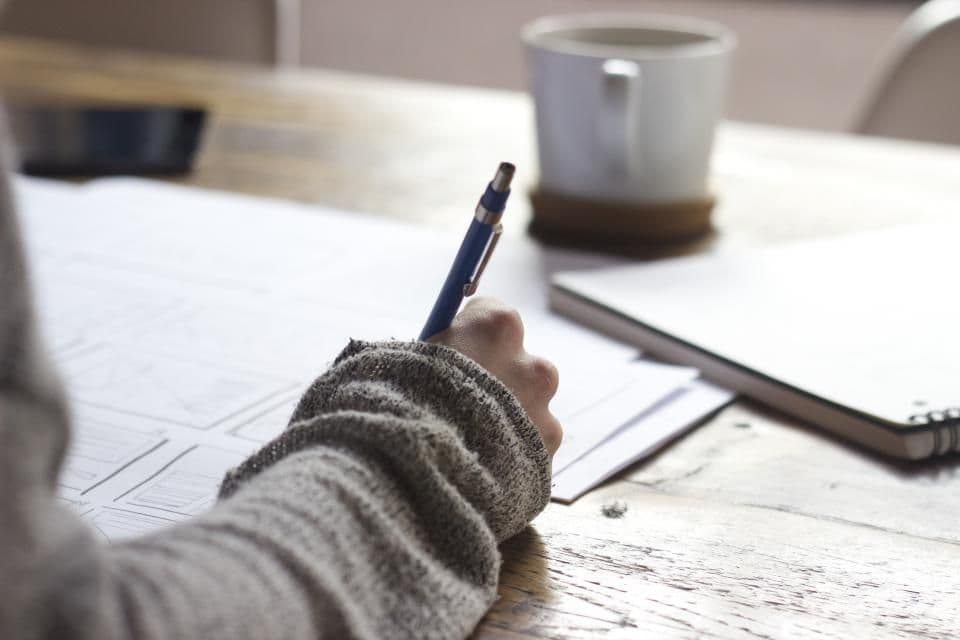 A hand writing on paperwork beside a cup of coffee